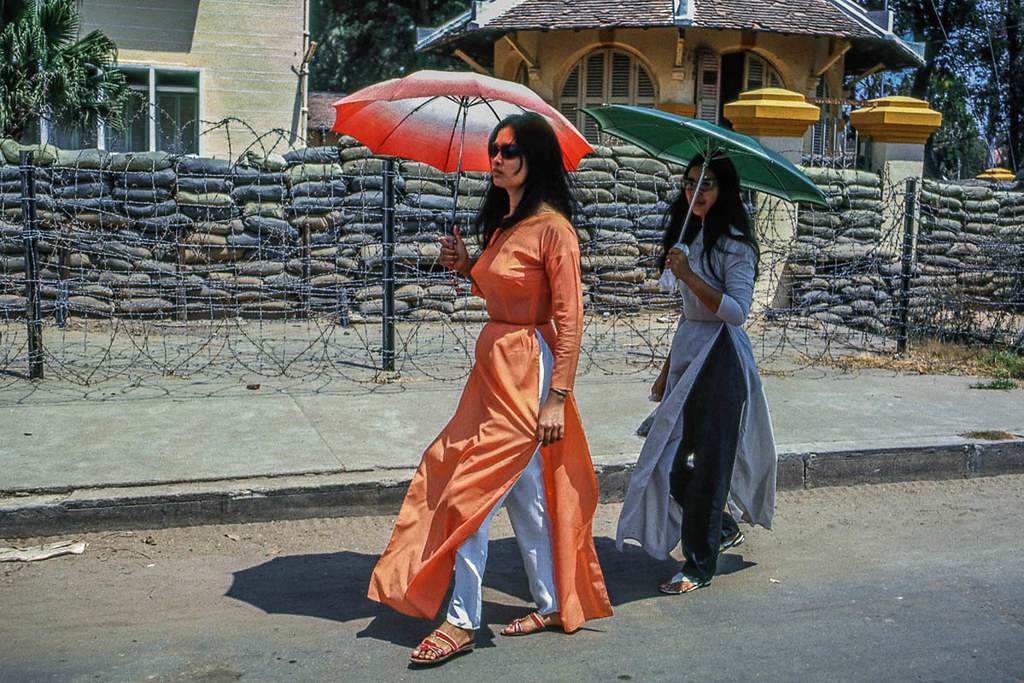 Hai phụ nữ trong trang phục áo dài đi ngang qua hàng rào kẽm gai và những bao cát. Ảnh chụp tại Mỹ Tho 1969
